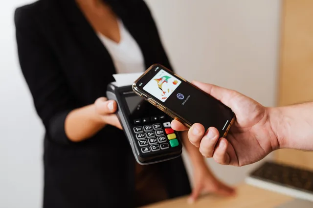 A person using a smartphone for contactless payment at a retail store counter. UGC creator payment processing fees: Stripe vs PayPal vs ACH